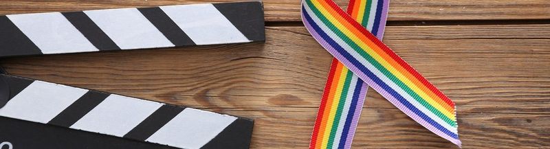 On the left is the black and white striped top of a clapperboard. Beside it is a Pride rainbow ribbon. They are both sat on a wooden table.